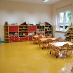 Zdjęcie przedstawia sale przedszkolną. Kolorowe szafki w kształcie parowozu. Żółta podłoga i beżowe ściany. Na pierwszym planie stoliki z krzesłami.