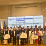 Uczniowie i nauczyciele z nagrodzonych szkół w Małopolsce pozują razem na scenie z dyplomami i symbolami nagród. Za nimi ekran z informacją o laureatach oraz flagi Polski i Unii Europejskiej.
