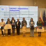 Laureaci konkursu edukacyjnego z województwa małopolskiego stoją na scenie z nagrodami i dyplomami. W tle widoczna jest lista nagrodzonych szkół oraz flagi Polski i Unii Europejskiej.