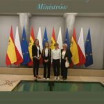 Cztery osoby stoją przed rzędem flag Polski i Unii Europejskiej oraz Hiszpanii. Na zdjęciu widnieje napis 