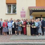 Grupa dyrektorów szkół i radnych Gminy Budzów stoi na zewnątrz przed budynkiem. Mężczyźni ubrani są w garnitury, a kobiety w sukienki lub kostiumy ze spodniami. Niektóre kobiety trzymają koszyki z roślinami i kwiatami. Na budynku widoczne są tablice z napisami „RADA GMINY W BUDZOWIE” oraz „URZĄD GMINY W BUDZOWIE”.