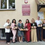 Grupa siedmiu osób, sześciu kobiet i jednego mężczyzny, stojąca na zewnątrz przed budynkiem. Wszystkie osoby na zdjęciu trzymają małe, drewniane koszyki z roślinami i kwiatami. Kobiety ubrane są w różnorodne stroje, a mężczyzna ma na sobie białą koszulę i ciemne spodnie. Widoczne są te same tablice na budynku. To zdjęcie przedstawia wyłącznie dyrektorów szkół Gminy Budzów.