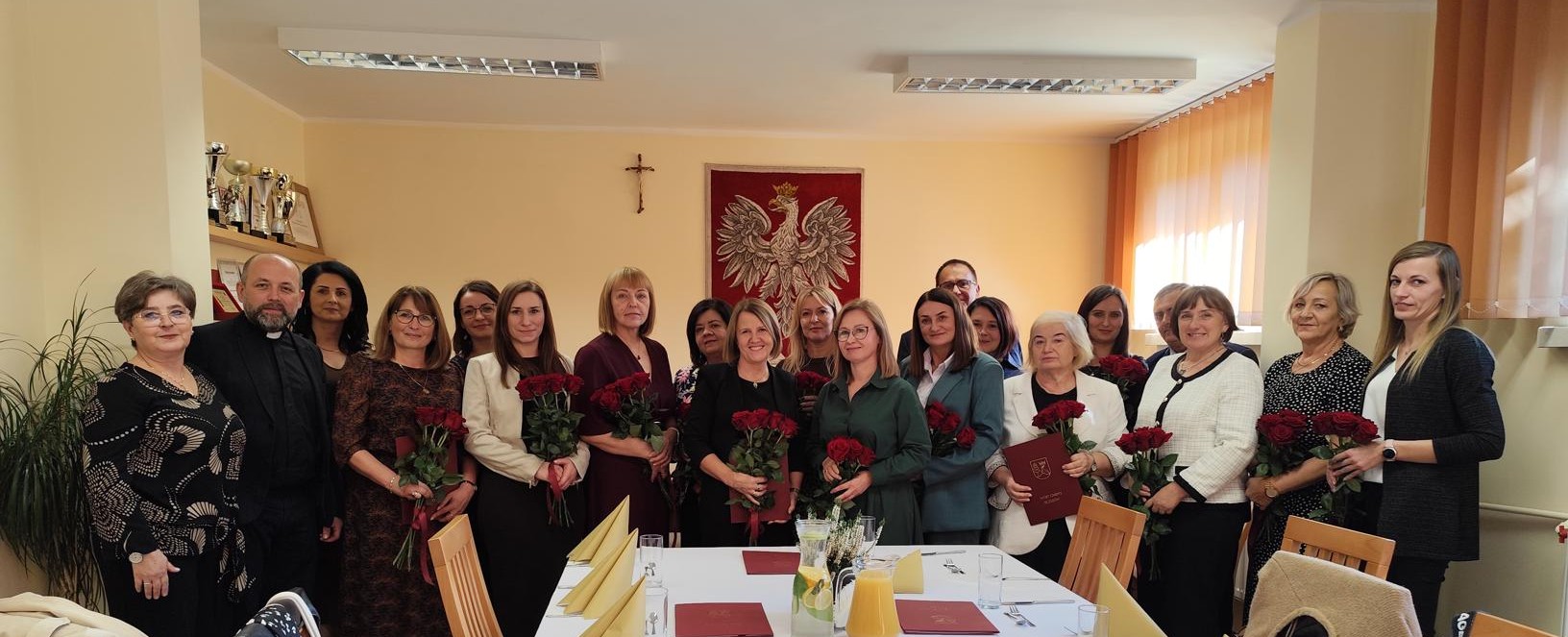 Grupa dyrektorów z bukietami róż stojąca pod polskim godłem. Uroczyste spotkanie w Urzędzie Gminy.