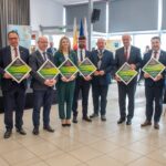 Grupa dziewięciu osób stoi w rzędzie w nowoczesnej sali. Większość z nich trzyma pamiątkowe tabliczki w kształcie rombu z napisem „ZNAK DOBREJ DROGI”. Uczestnicy są ubrani w stroje wizytowe – garnitury, marynarki i eleganckie komplety. W tle widoczne są flagi oraz baner z herbem Gminy Siepraw.