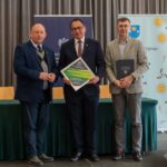 Trzech mężczyzn stoi na tle zielonego stołu i banerów informacyjnych. Środkowy mężczyzna trzyma pamiątkową tabliczkę „ZNAK DOBREJ DROGI DLA GMINY BUDZÓW”. Osoba po prawej trzyma ciemną teczkę z napisem „Wojewoda Małopolski”. Wszyscy są uśmiechnięci i patrzą w stronę obiektywu.