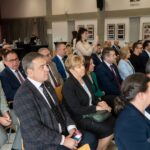 Ujęcie z boku na publiczność siedzącą w rzędach podczas oficjalnego spotkania. W kadrze widać profil i plecy osób w strojach biznesowych, skupionych na wydarzeniu. W tle kobieta robi zdjęcie telefonem, a na ścianach wiszą oprawione fotografie.