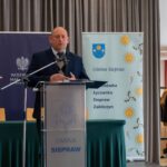 Mężczyzna w ciemnej marynarce i swetrze przemawia przy przezroczystej mównicy z napisem „GMINA SIEPRAW”. Za nim znajdują się zasłony oraz dwa banery: jeden z godłem Polski (Wojewoda Małopolski), a drugi z herbem i nazwami miejscowości gminy Siepraw.